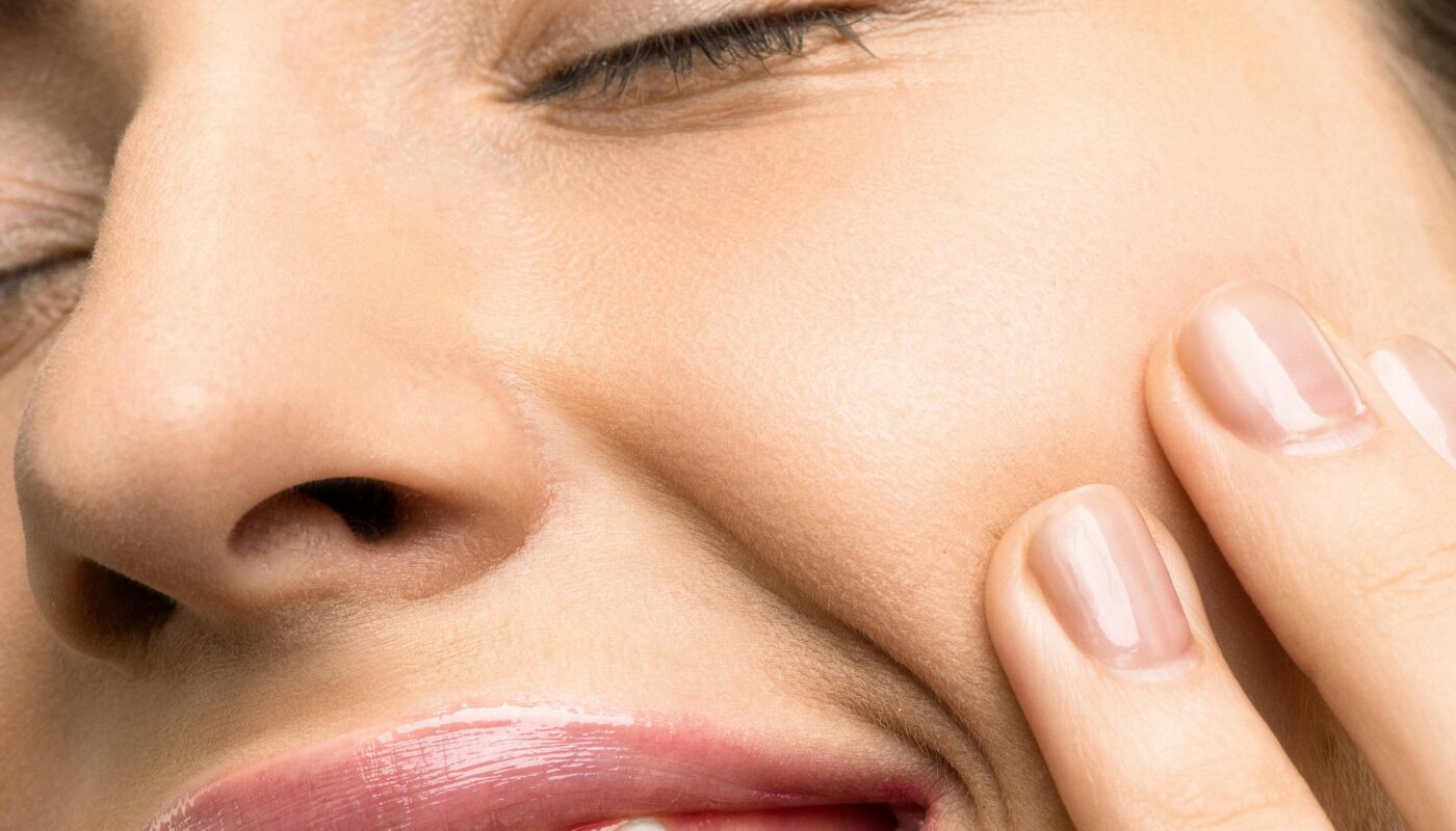 A close-up portrait of a joyful woman with closed eyes, pink lipstick, and her hand touching her cheek.