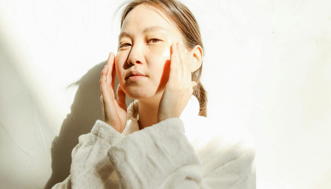 Woman in bathrobe applying skincare with natural light and shadows.