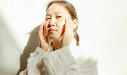 Woman in bathrobe applying skincare with natural light and shadows.