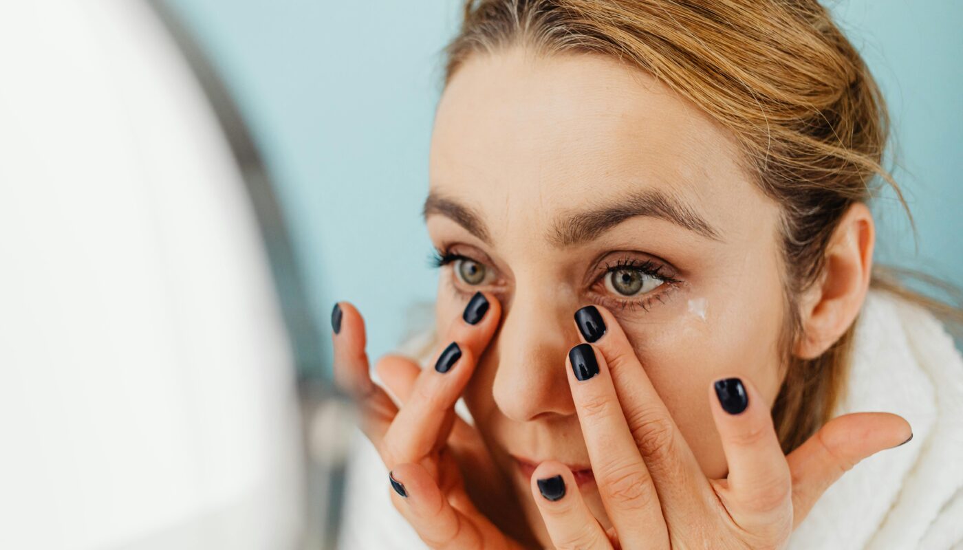 Close-up of a woman applying facial cream in front of a mirror with blue background.