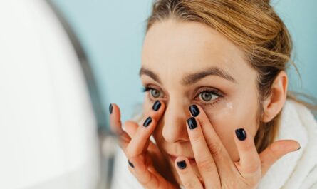 Close-up of a woman applying facial cream in front of a mirror with blue background.